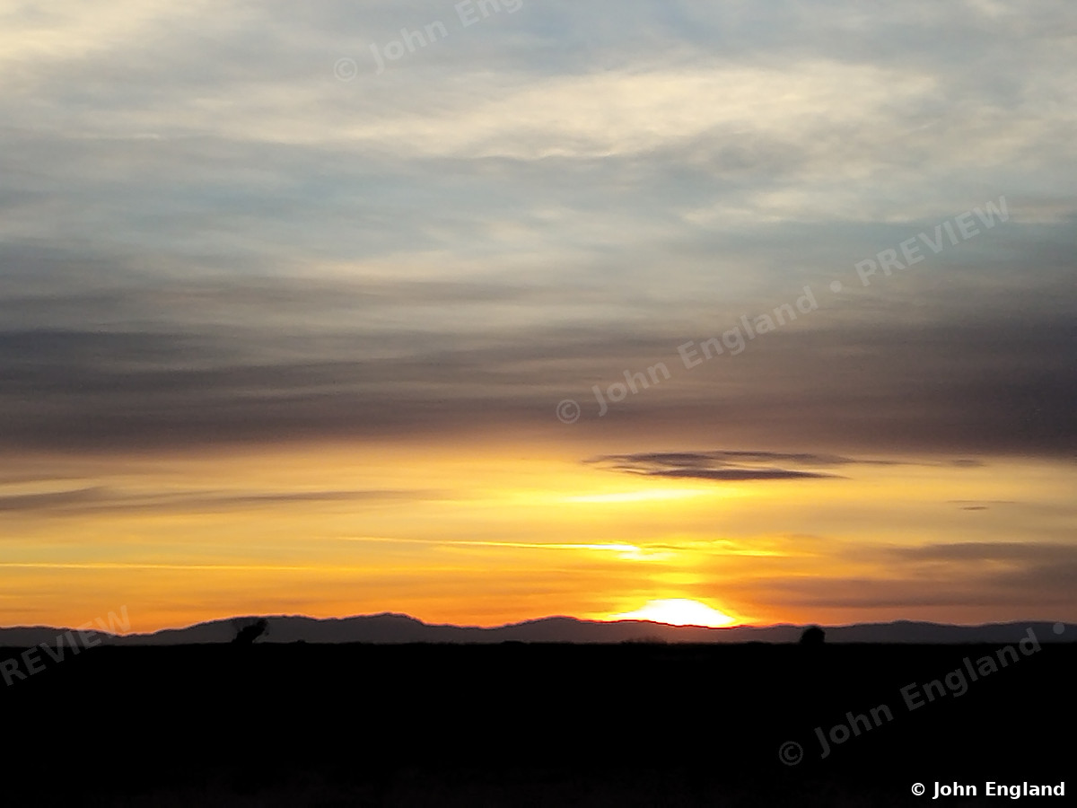 Ridge Sunset – Sun sinking behind a distant ridge under layered clouds.