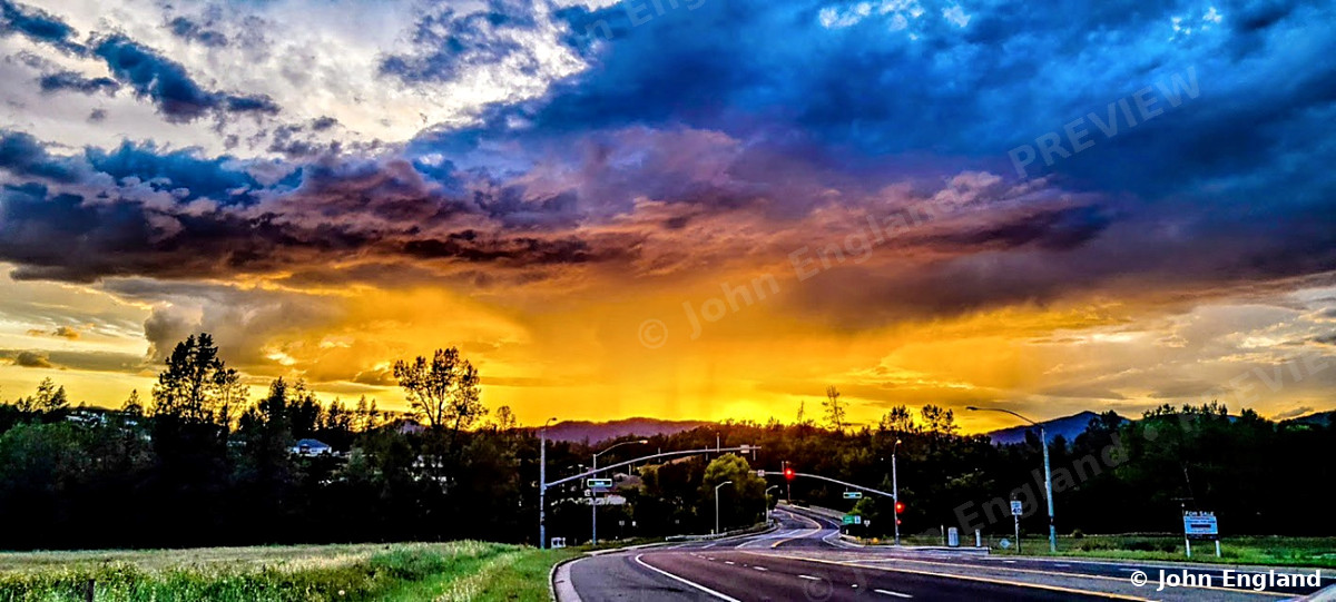 Storm Sunset over the Road – Dramatic storm clouds and orange sunset over a roadside tree line.