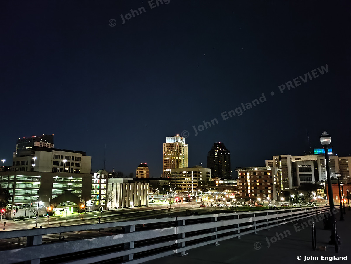 Sacramento Skyline After Dark – Downtown Sacramento skyline at night from a riverfront walkway, Sacramento, CA. Feb 2021. John England.