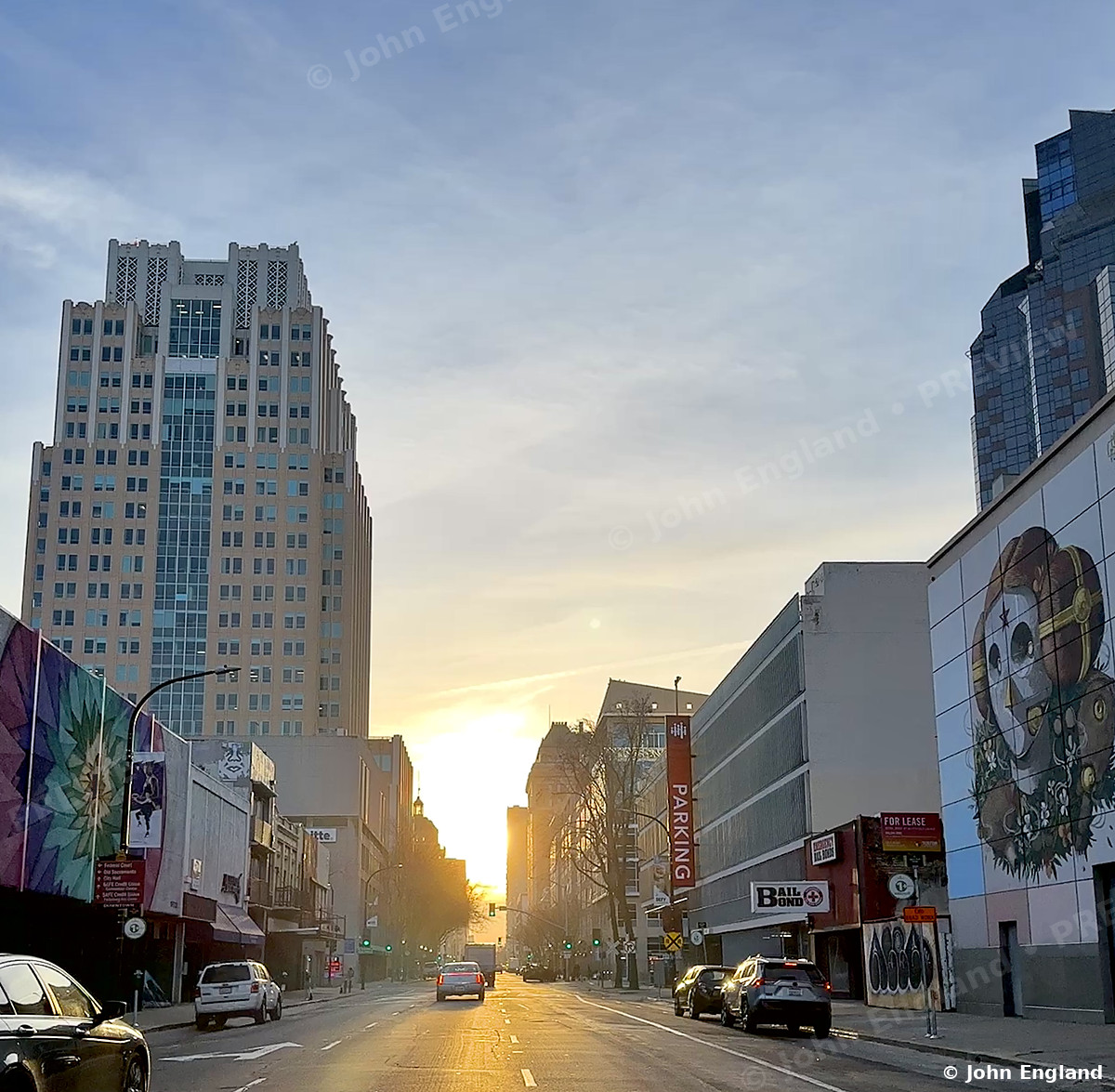 Sunrise Down a Downtown Street – Sunrise light down a downtown Sacramento street lined with towers and a mural, CA. Feb 2023. John England.