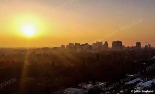 Golden Sunrise over the City – Aerial sunrise with sun flare over trees and distant skyline.