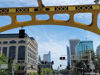 Under the Yellow Bridge Truss – Looking up through a yellow bridge truss toward downtown Sacramento towers, Sacramento, CA. Mar 2022. John England.